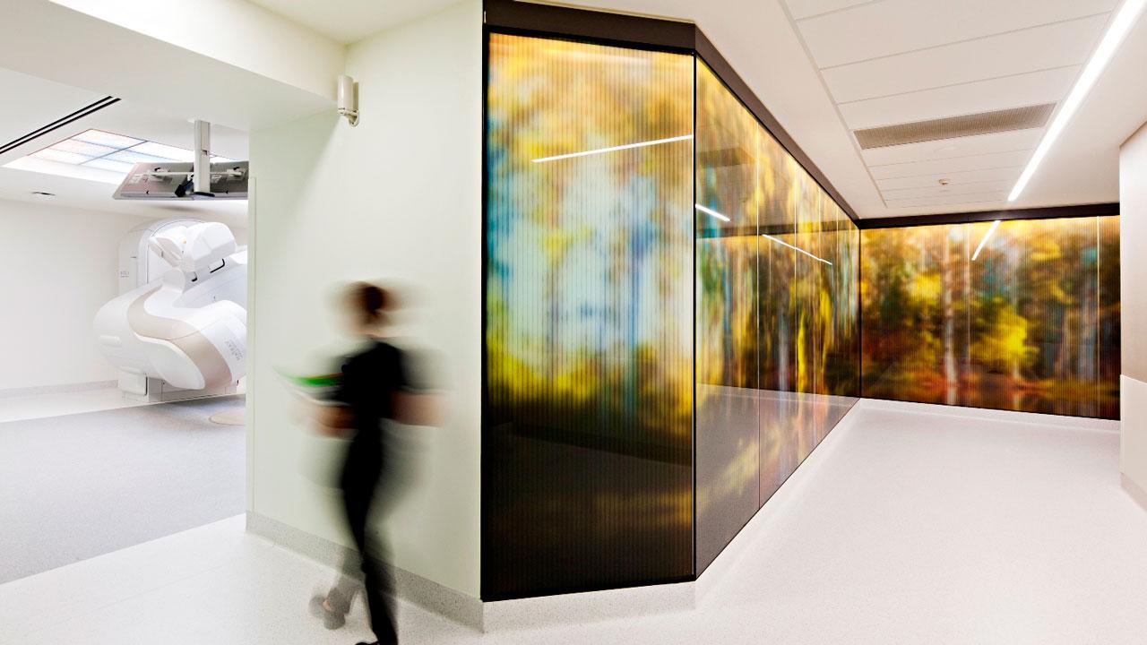 A medical professional walking up a corridor with a colored glass wall towards an open CT scanning room