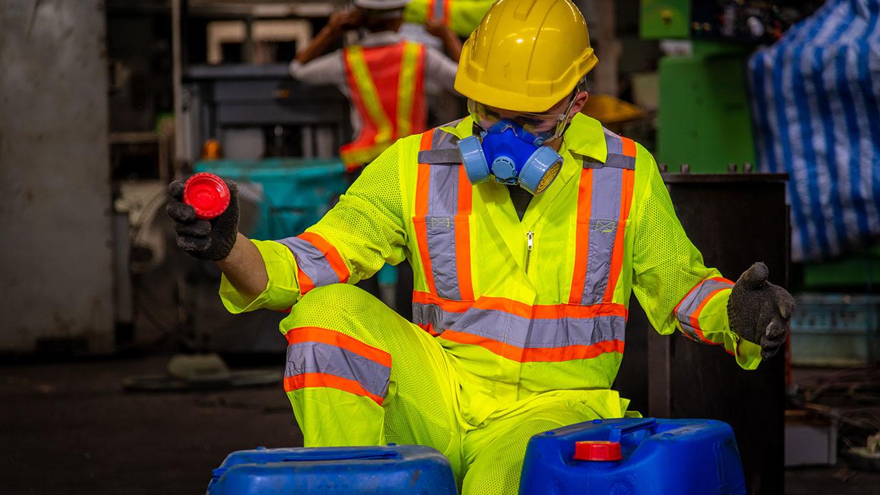 An engineer wearing safety uniform, black gloves, gas mask, and hard hat checking chemical tank in a factory